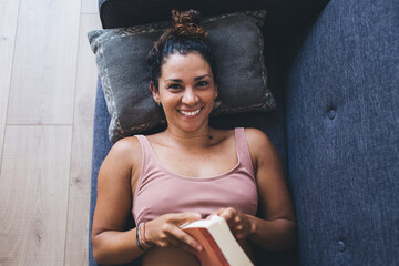 Cheerful Caucasian female holding book and laughing at funny story enjoying spending free time at...
