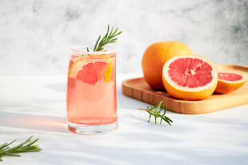 Elegant glass with a fresh, cooling cocktail of fresh grapefruit or red orange with ice cubes and a sprig of rosemary on a gray background on a bright sunny day. Vacation and summer drinks concept.