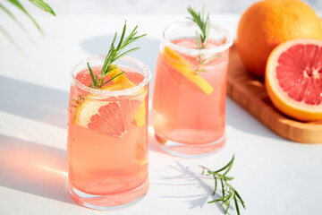 Two glasses with a fresh, cold summer cocktail of grapefruit and ice, garnished with a sprig of rosemary and fresh fruit slices. Red orange drink. Shadows on a sunny day.