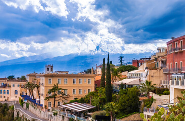 Naklejka na ściany i meble Taormina, Sicily, Italy. Panoramic view over Taormina town on hilltop and Etna mount volcano among clouds in blue sky. Popular tourist destination