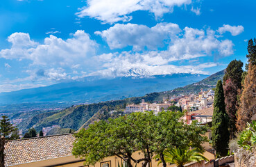 Obraz premium Taormina, Sicily, Italy. Panoramic view over Taormina town on hilltop and Etna mount volcano among clouds on blue sky. Popular tourist destination