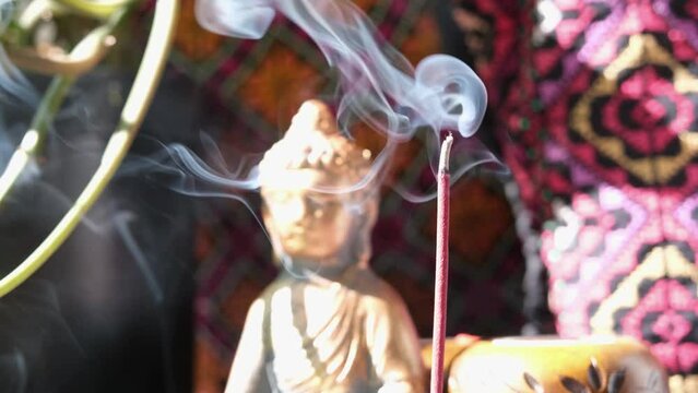 An incense stick burning and generating aromatic smoke surrounded by Buddha statue and eastern ornament pillows