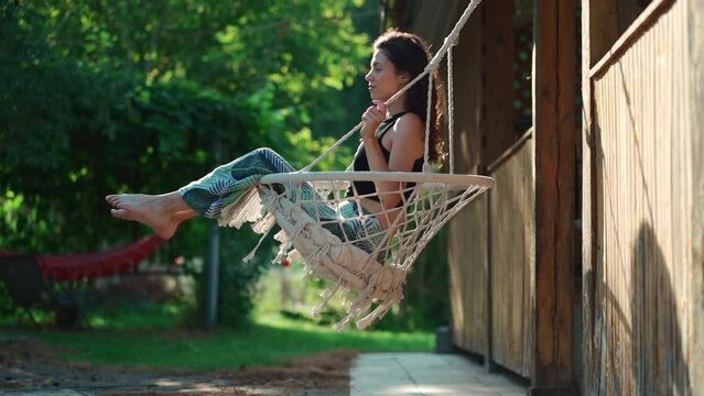happy young female riding on macrame swing chair near country house outdoors