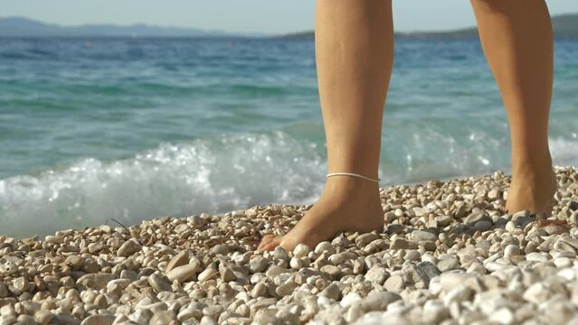 SLOW MOTION, CLOSE UP: Young lady walking along pebbly seashore with bare feet. It is a sunny day and she is on a relaxing holiday walk along beautiful Dalmatian beach by crystal clear Adriatic Sea.
