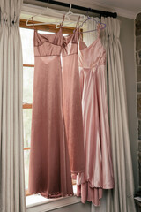 Three long pink bridesmaid dresses hang against the window. Beautiful bridesmaid dresses. Stylish dresses hang in the room and are ready for the wedding ceremony