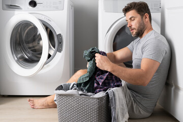 Man doing laundry at home near washing machine. Husband man doing laundry at home. Sexy guy washer putting clothes in the washing machine. Household concept. Washer and dryer, laundromat. - obrazy, fototapety, plakaty