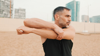 Naklejka na ściany i meble Athletic mature man stretches arm muscles while standing on the beach in the morning