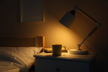 Stylish modern desk lamp, books and cup of drink on white nightstand in dark bedroom