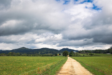 Perspective of gravel roadway through fields of green grass with a range of hills in the background, all under an overcast and threatening sky.