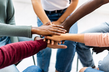 Fototapeta na wymiar Young group of people sitting in circle stacking hands. Multiracial friends putting their hands together showing unity, support and community.