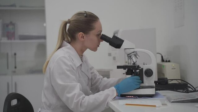 Female medical professional works with samples and analyzes in medical laboratory behind microscope. Woman lab technician analyzing sample through microscope in hospital. Medicine, microbiology.
