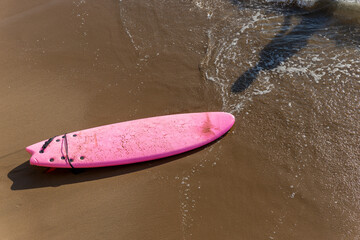 Pink surfboard on orange sea sand, splashed with sea water and a wave about to break over it.