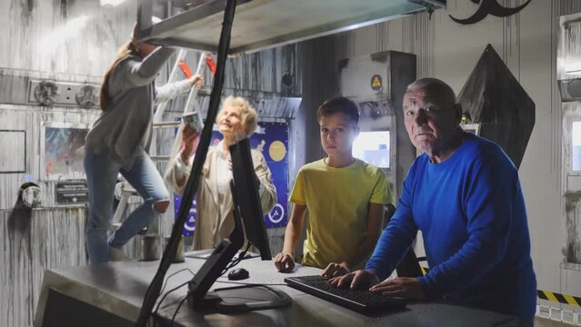 Young Boy And His Grandfather Using Computer To Solve Conundrum In Escape Room. Grandmother Helping Teenager Girl Who Standing On Stepladder In Background. 