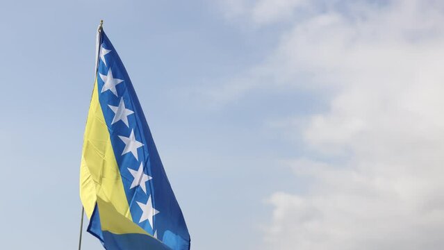 Large flag of Bosnia and Herzegovina fixed on metal stick waving against background of cloudy sky during daytime . High quality 4k footage