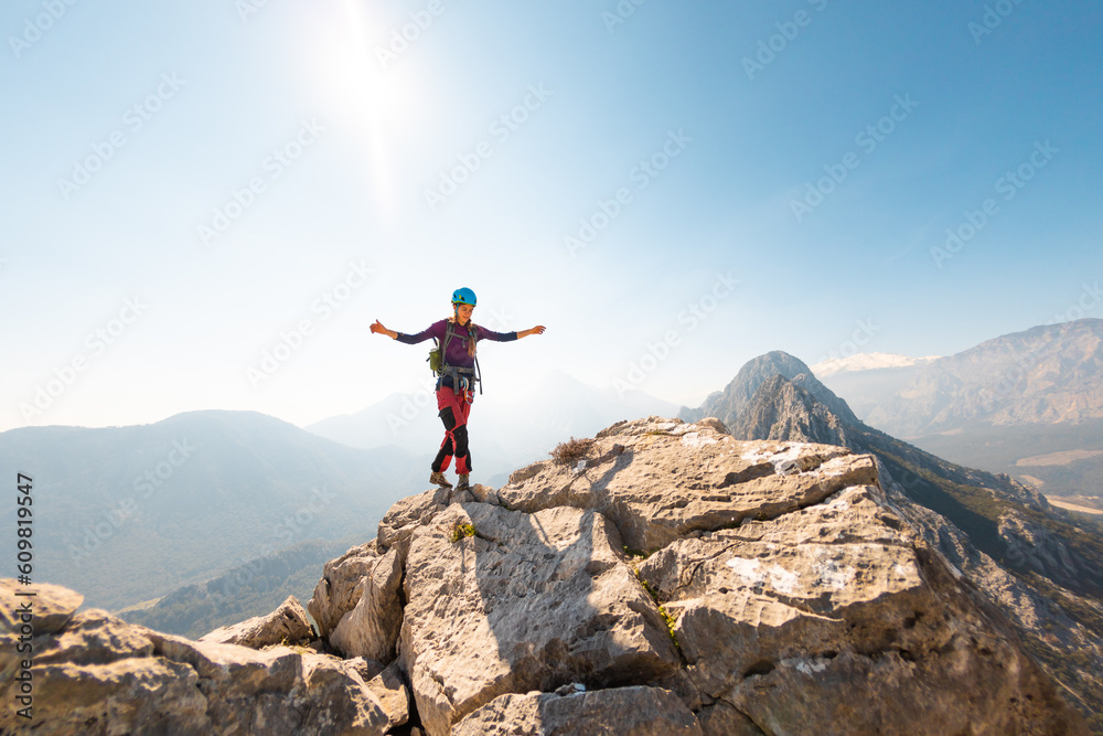 Wall mural young girl climber in a helmet and with a backpack stands on top of a mountain. mountain climbing