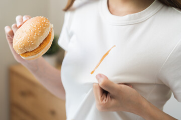 Cloth stain, disappointment asian young woman, girl eat food hand hold hamburger, burger show making tomato sauce smudged spilling, drop on white t-shirt, spot dirty or smudge on clothes, dirt stains.