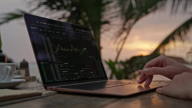 Female crypto trader at laptop checking candlestick charts online working remotely at outdoor tropical seaside cafe. Woman broker analyses exchange rates and bets on crypto stock by ocean closeup