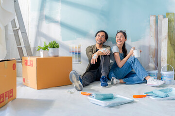 Asian Cheerful young marry couple having coffee break sitting among paint tools and accessories during the break in new apartment,home relocation renovate interior concept,happiness couple paint wall