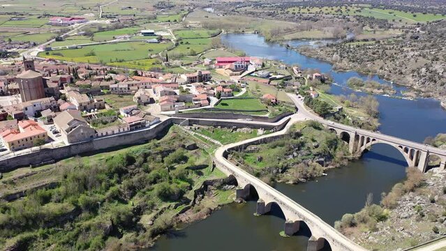 Drone view of Ledesma townscape on green hilly banks of Tormes river with medieval arched bridge in spring overlooking massive Hispano-Flemish Gothic building of church Santa Maria la Mayor, Spain 