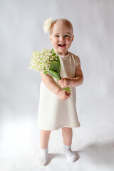 Portrait of a smiling little girl in a white dress on white background full-length. Toddler embraces a bouquet of fresh lilies of the valley . Gift for the holiday, the concept of purity, spring time