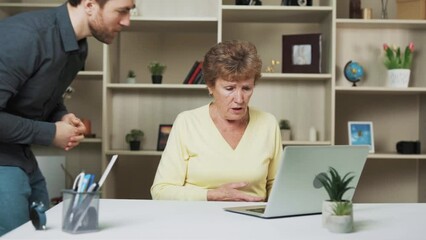 A woman is sitting at a table with a laptop in front of her. She is working on the laptop. Being older, she is having trouble with her laptop and asked a young man for help. He resolves her issue. - Powered by Adobe