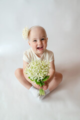 Portrait of a smiling little girl in a white dress on white background. Toddler embraces a bouquet of fresh lilies of the valley . Gift for the holiday, the concept of purity, spring time. 