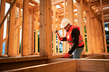 Carpenter constructing wooden frame house. Bearded man with glasses is hammering nails into the structure, wearing protective helmet and construction vest. Concept of modern ecological construction.