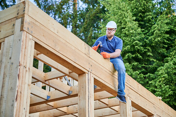 Carpenter constructing double-story wooden-framed house beside the woodland. Bearded man with glasses using hammer to drive nail while wearing safety helmet. Idea of contemporary ecological building.