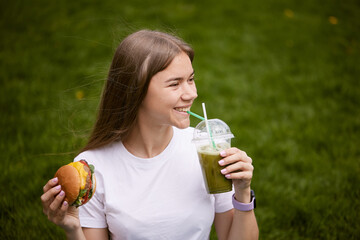 a young girl takes out a burger from a paper bag sitting on the green grass, the concept of food delivery, street food