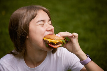 a young girl takes out a burger from a paper bag sitting on the green grass, the concept of food delivery, street food