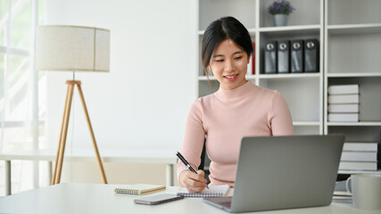A beautiful Asian businesswoman focuses on her work, taking notes and using her laptop