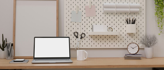 A laptop white screen mockup on a wooden table against the wall with white pegboard.