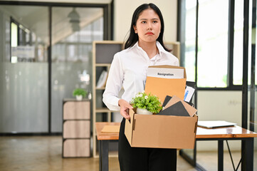 Sad and upset Asian female office worker stands in the office with a cardboard box with her stuff