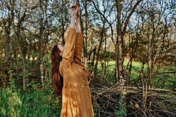 a beautiful, slender woman with long hair walks in the shade near the trees, dressed in a long orange dress, enjoying the weather and the weekend, joyfully raising her hands up