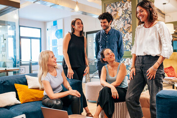 Team of young positive coworkers gathering around table with laptop and discussing ideas for startup project while having meeting in creative workspace.