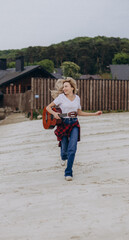 Dynamic energetic girl in a white t-shirt and jeans holds a guitar in her hands, running along the lakeside, looking sideways and smiling. Positive cheerful woman is enjoying outdoor leisure.
