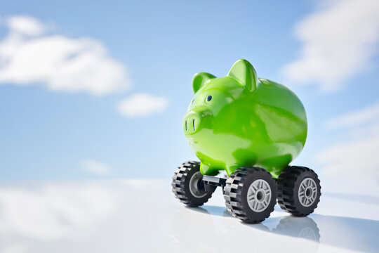 Green piggy bank money box on wheels against blue sky background, new vehicle purchase, insurance or driving and motoring cost