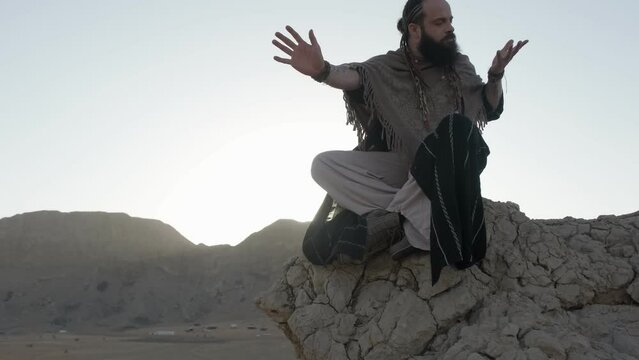 A youthful young shaman in the lotus position prays on a rock against the backdrop of sunset among the sand dunes of the desert