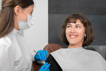Young smiling woman is sitting in a dental chair and looking at a dentist who is holding dental instruments in his hands to check the oral cavity
