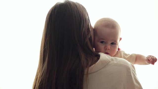 Brown-haired mother hugs baby daughter demonstrating strong parent love in bedroom. Happy parent takes care of infant girl in light room