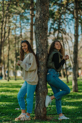 Long haired brunette girl burst out laughing while having a fun conversation with her lovely curly haired friend. They are both holding a flower in their hands and leaning against a tree in the park