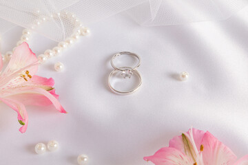 Luxurious wedding background design. Two white gold engagement rings with diamonds on white satin fabric with pearls and delicate pink flowers.