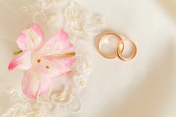 Two gold engagement rings and pink astromeria flower on a beige satin background. Wedding background. A copy space.
