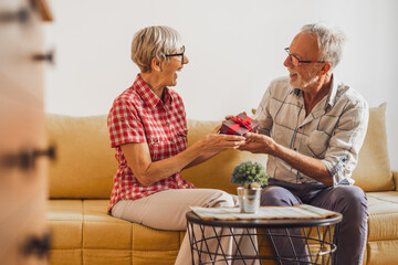 Mature couple sitting in living room and celebrating their anniversary. Husband is giving present to his wife.