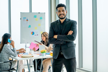 Portrait of Asian business man stand with arm-crossed and look at camera show confidence in front of other co-workers discuss on the back in office.