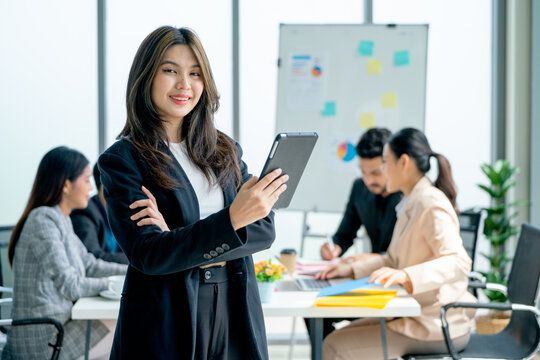 Portrait of young Asian business woman hold tablet and stand with look at camera in front of other co-workers discuss on the back in office.