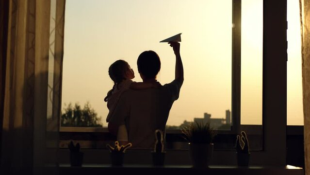 Mom imitates airplane flight using a paper plane while daughter watches her from the window during sunset.