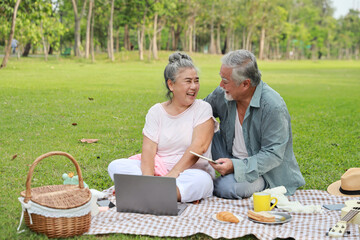 Happy asian senior man and woman sitting on blanket and having fun on picnic together in garden outdoor. Lover couple using computer and writing at the park. Happiness marriage lifestyle concept.