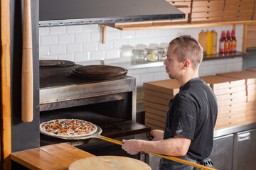 The chef prepares pizza. Raw pizza ready to bake. Cook in a blue apron in the kitchen. with a shovel in his hands. boxes for food delivery on background.