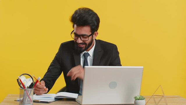 Thoughtful journalist indian businessman making notes, writing down thoughts with pen into notepad notebook diary, to do list, good idea at office workplace desk. Hindu man in suit working on laptop